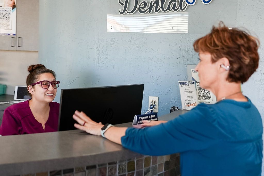Two people talking at a dental office reception desk, one smiling behind the counter.