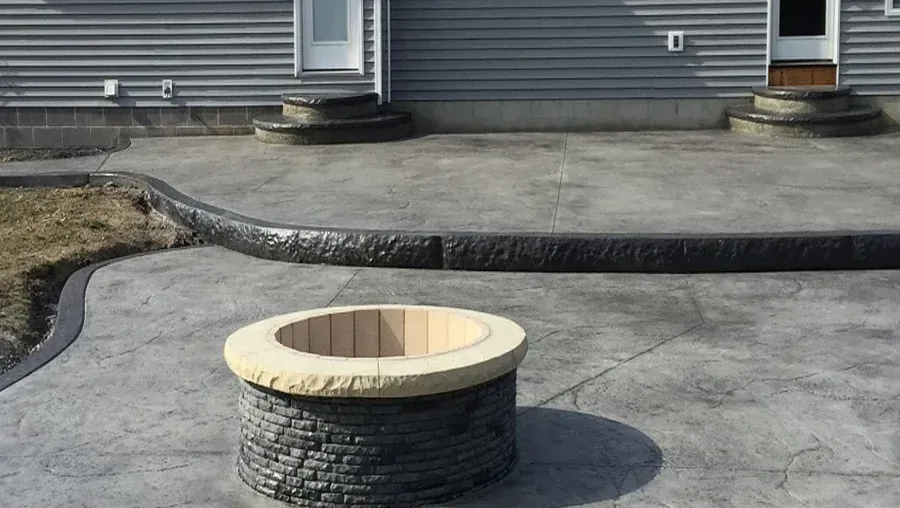 Decorative concrete patio with a round stone fire pit in front of a house's gray siding