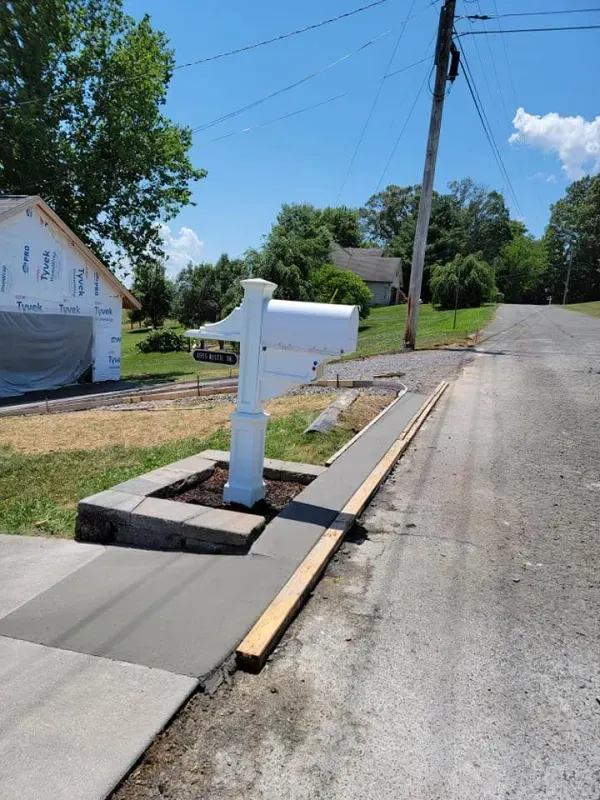 White mailbox on a concrete pad beside a rural road with a house and trees in the background