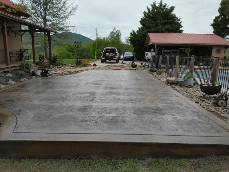 Fresh concrete patio in a rural yard, with houses, potted plants, and parked vehicles in the background.
