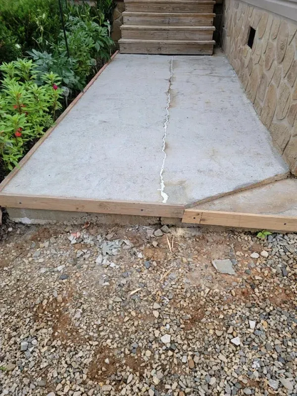 Cracked concrete walkway leading to steps, with gravel at the base and stone wall on the right.