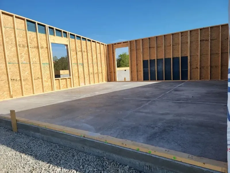 Unfinished wooden-framed building interior with concrete slab floor and open window and door openings.