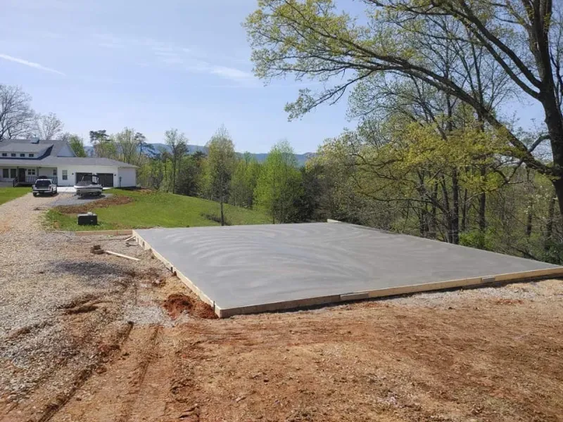 Concrete slab in a cleared wooded lot beside a house and gravel driveway under a sunny sky