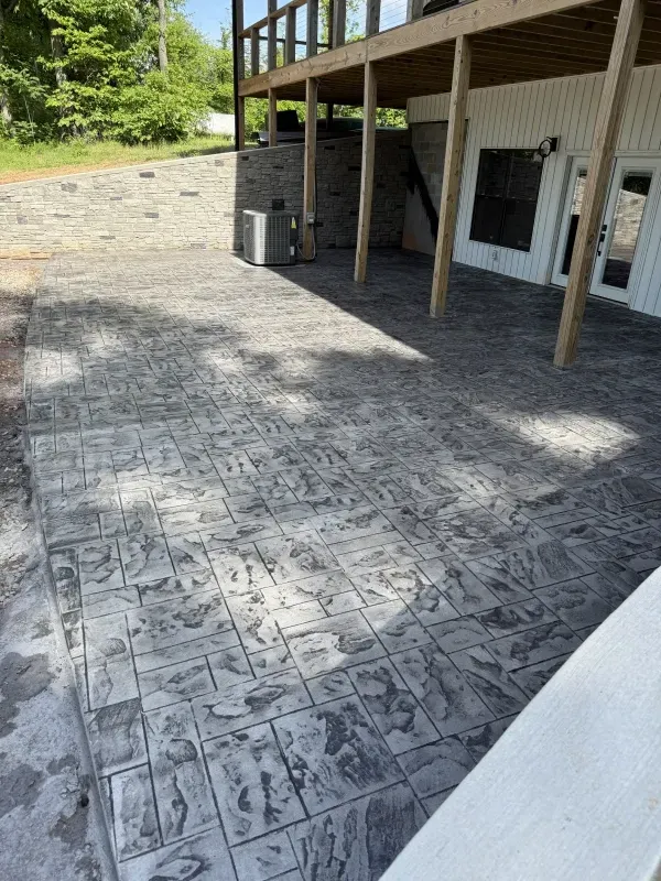 Stamped concrete patio beside a house with support posts and a sloped gravel yard nearby