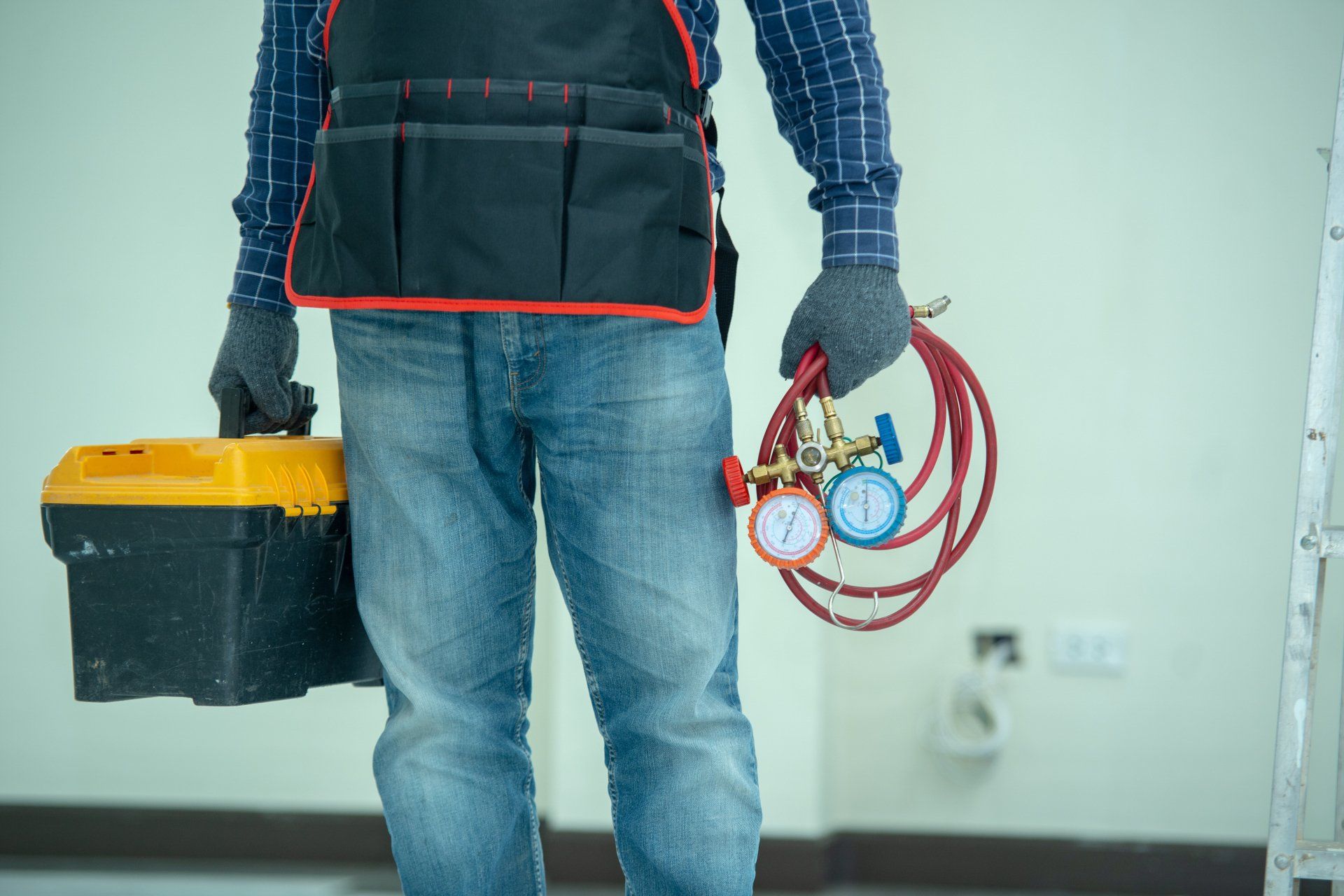 Un technicien en CVC, portant des gants de travail et un jean, tient une boîte à outils et des manomètres dans un environnement intérieur.