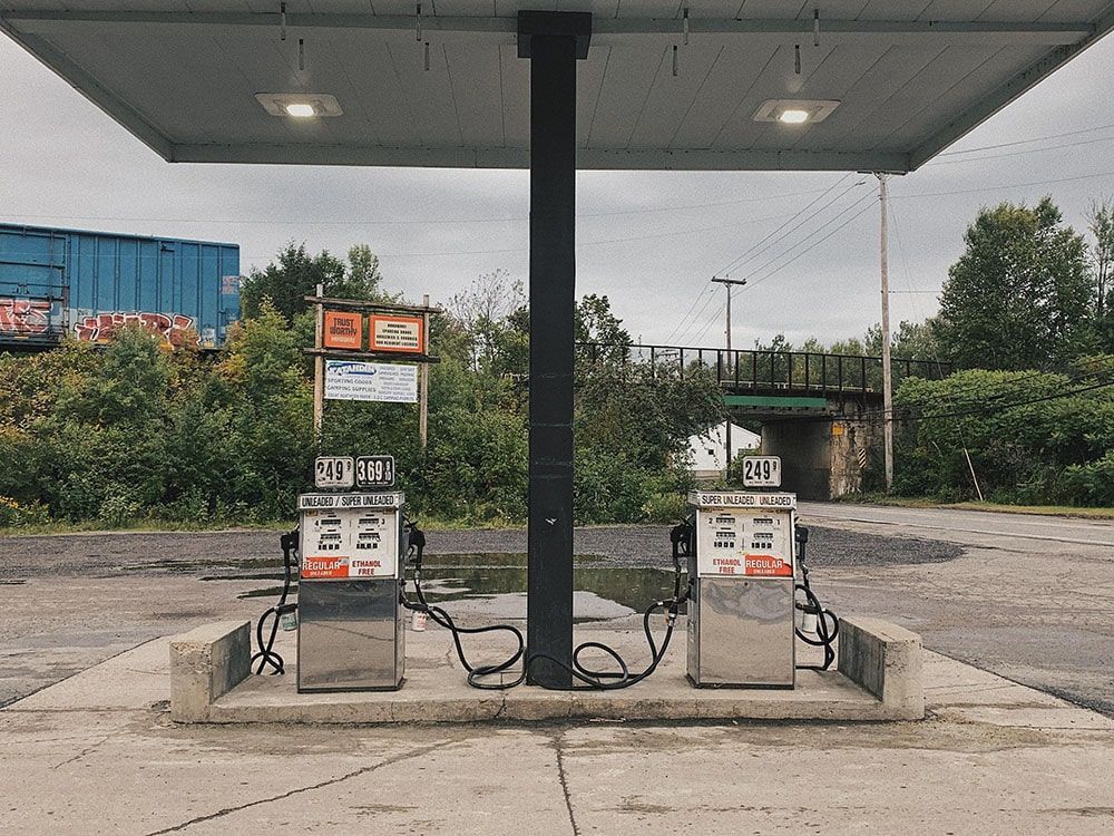 Two old gas pumps under a canopy at an abandoned roadside station, with weeds and containers nearby.