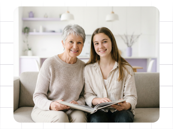 Two women sit on a couch, smiling and reading a book in a bright living room.