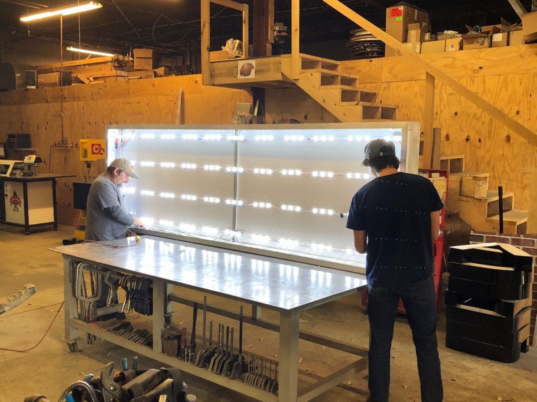 Two people working on a large light fixture in a workshop; white lights on the front.