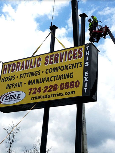 Hydraulic services sign being installed, two workers in lift, yellow and black sign, clear blue sky.