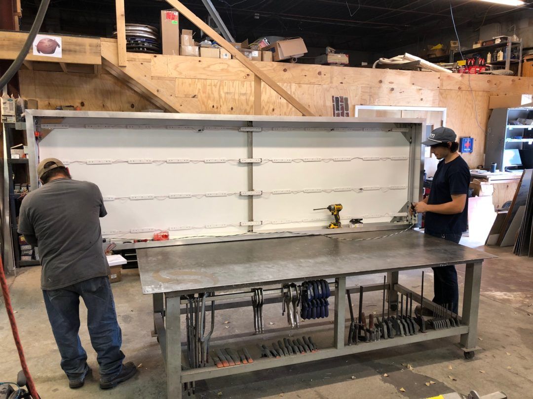 Two people working on a metal frame, possibly a workbench, in a workshop.