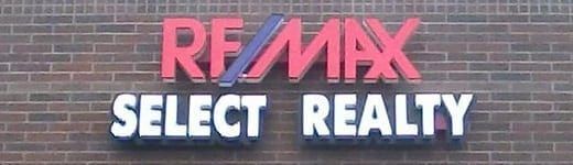 RE/MAX Select Realty sign on a brick wall. The RE/MAX text is red with a blue and purple line; the text 