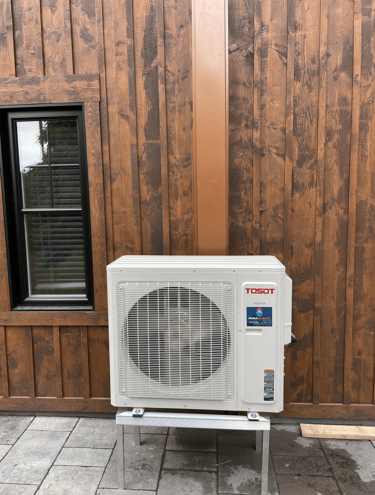Unité de climatisation blanche sur un support métallique, adossée à un bâtiment en bois brun.