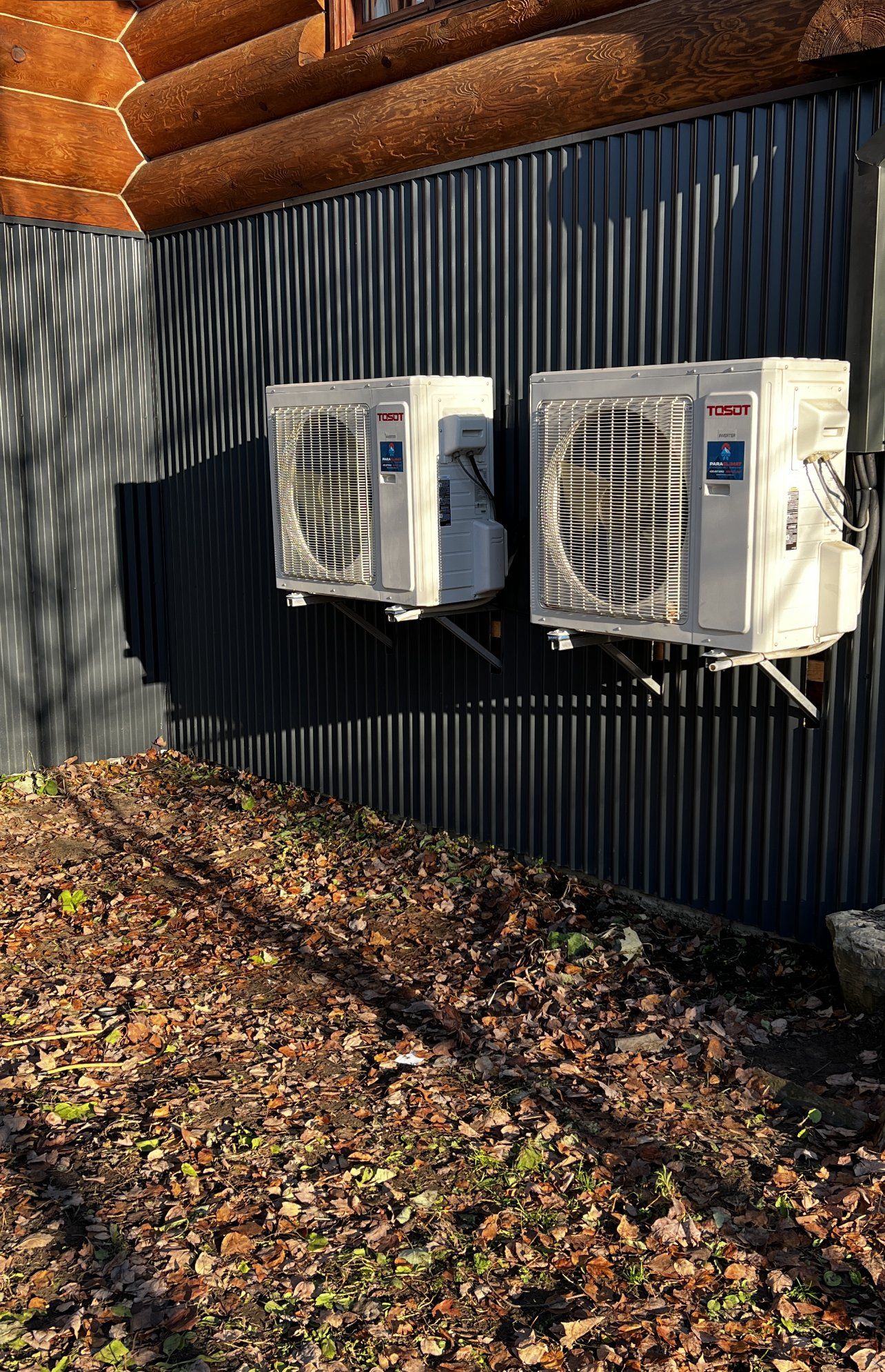 Deux unités de climatisation blanches fixées sur un mur en tôle ondulée sombre, près d'une cabane en rondins.