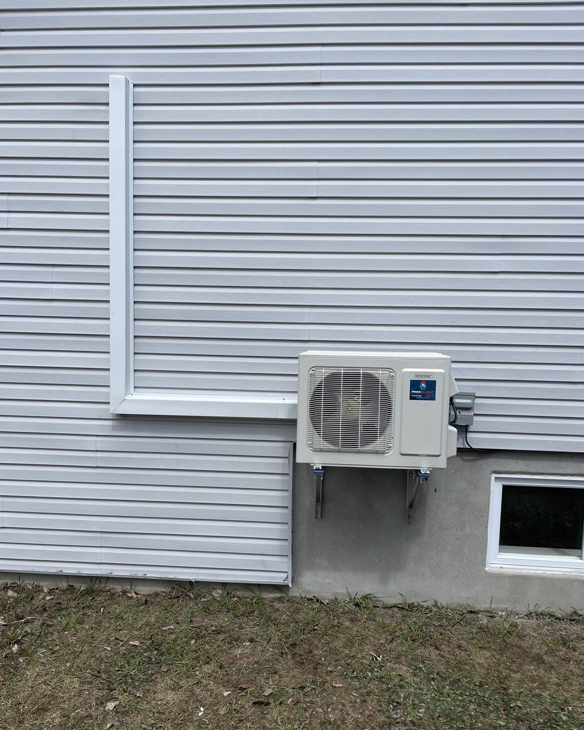 Unité de climatisation fixée sur un mur en béton, sous une fenêtre et un revêtement extérieur.
