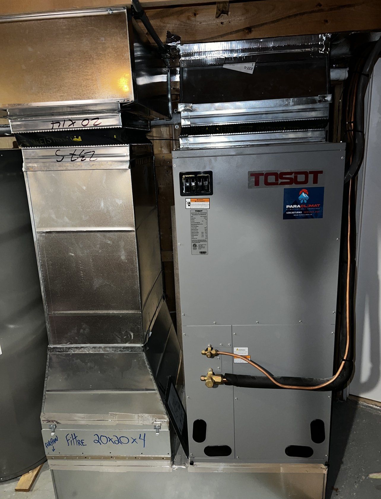 Unité de climatisation TOSOT avec conduits d'aération dans un sous-sol ; tuyauterie en métal argenté et en cuivre visible.