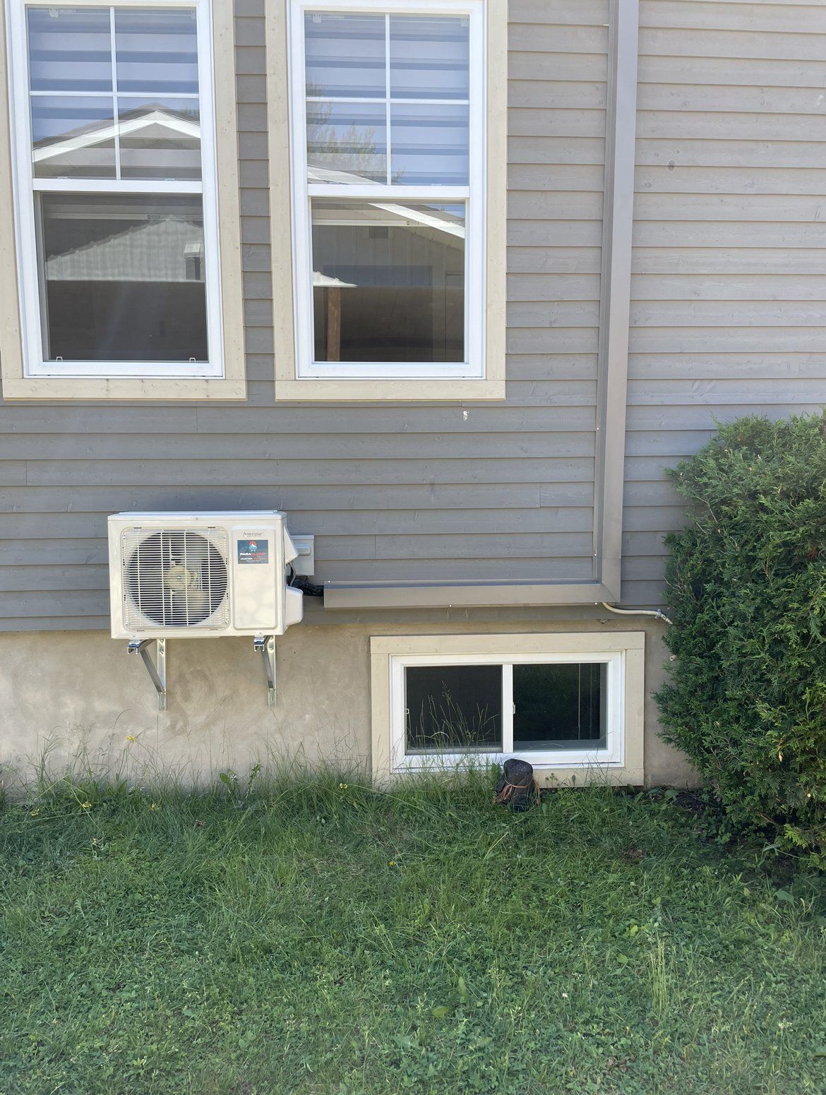 Extérieur d'une maison avec deux fenêtres et un climatiseur fixé sur un mur à bardage gris. Une petite fenêtre se trouve en dessous.