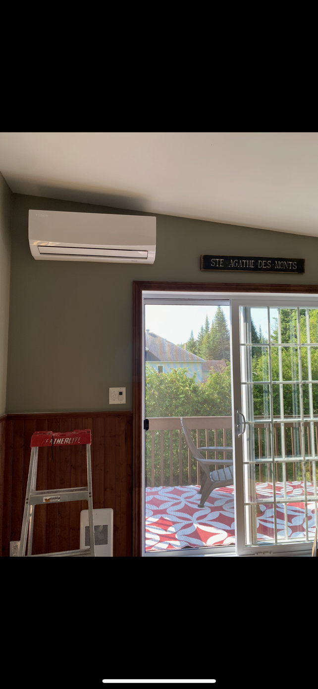 Un climatiseur intérieur est installé sur un mur vert au-dessus d'une porte coulissante en verre. Une échelle rouge se trouve près de la porte.
