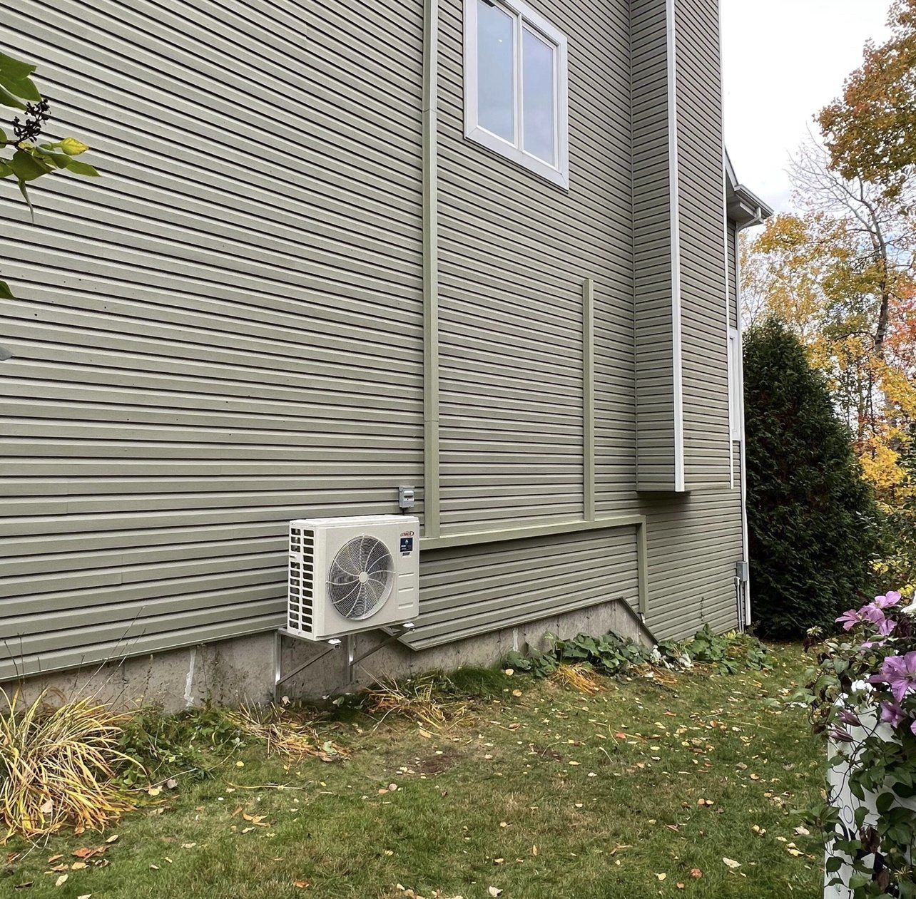 Un climatiseur extérieur monté sur le mur extérieur d'une maison au revêtement vert, entouré d'herbe et de feuillage.