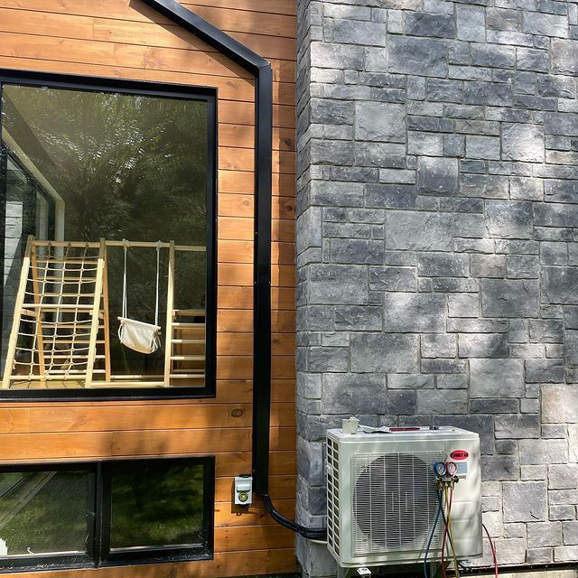 Extérieur d'une maison avec un climatiseur adossé à un mur en pierre et un tuyau noir remontant le long du bardage en bois.