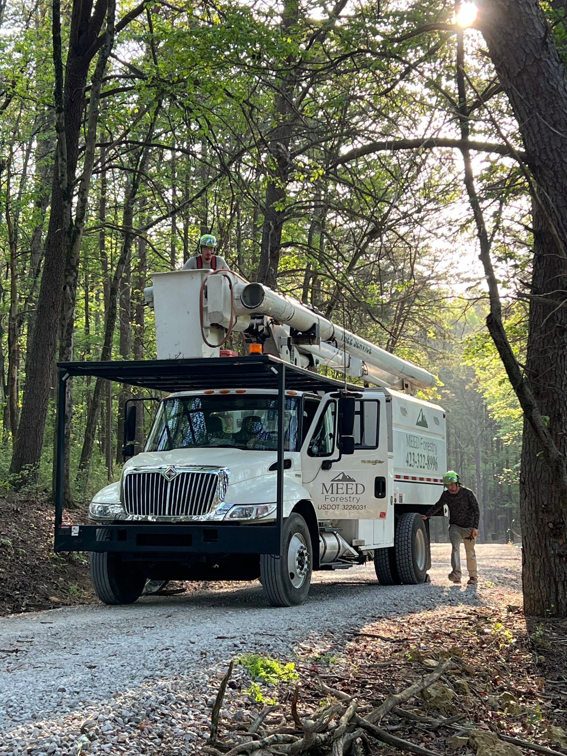 A utility bucket truck parked on a gravel road in a wooded area, with one worker in the bucket and another standing nearby.