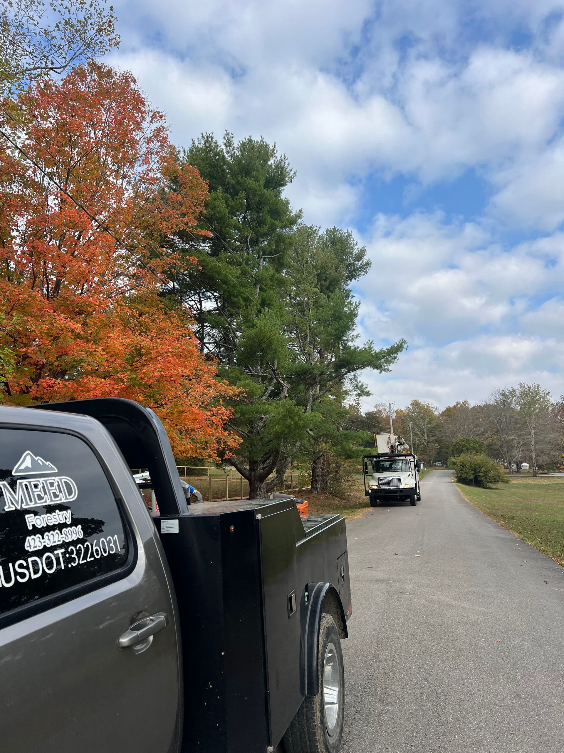 A gray utility truck parked on a rural road with a white bucket truck in the distance under a blue sky with autumn trees.