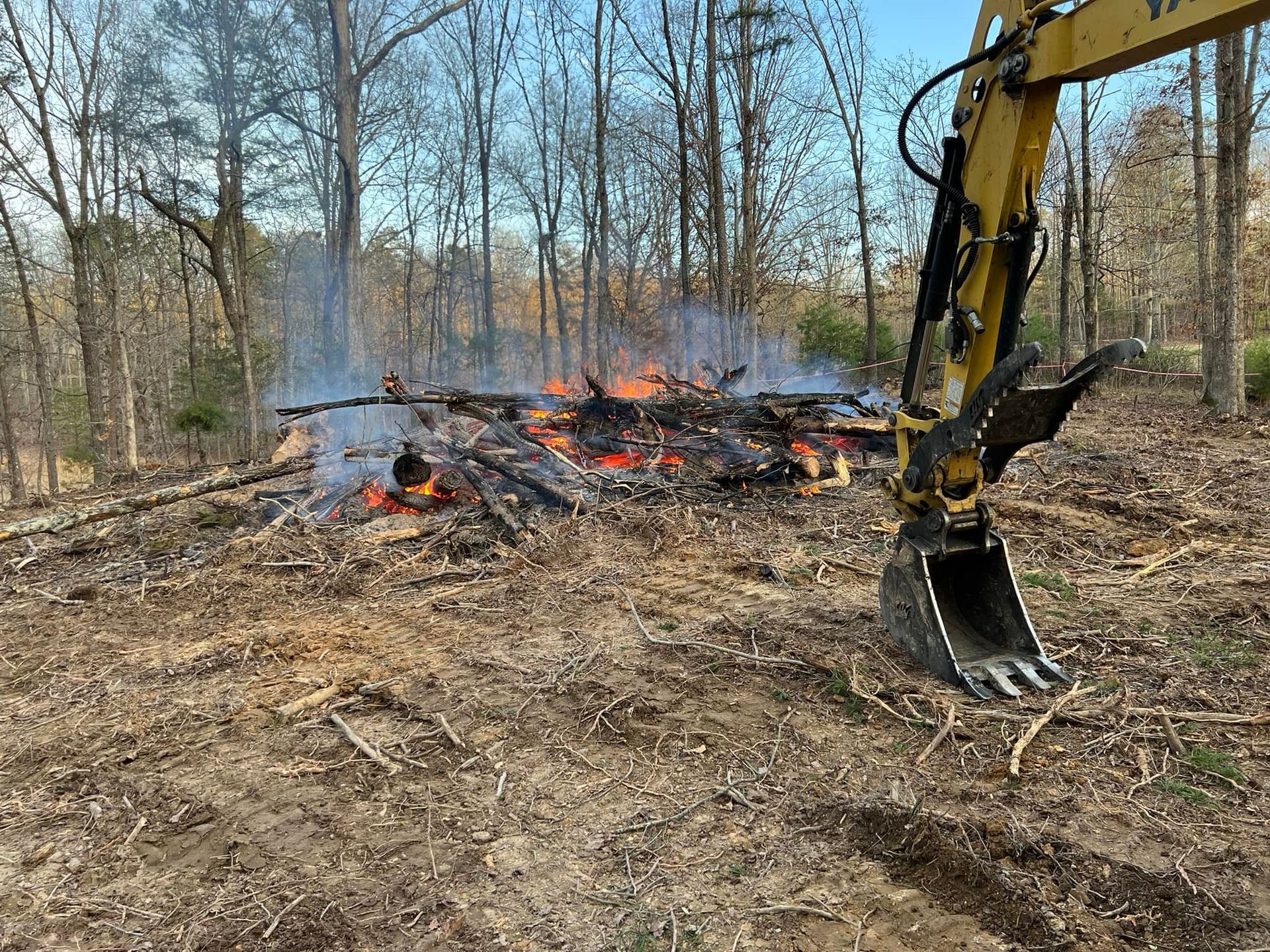 A yellow excavator arm stands beside a brush pile burning in a cleared, wooded lot.