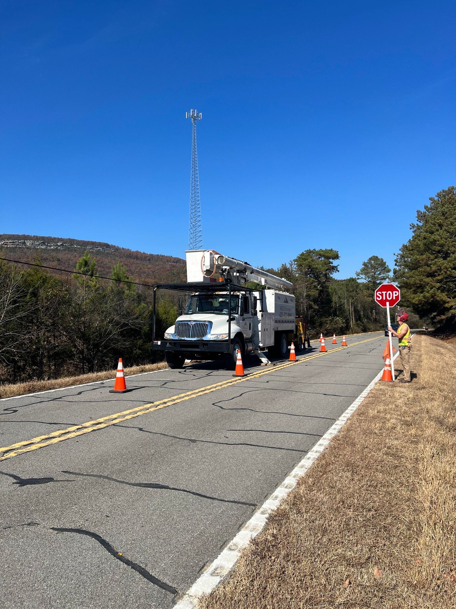 A utility truck with an extended mast blocks a rural road, with traffic cones and a flagger directing traffic nearby.