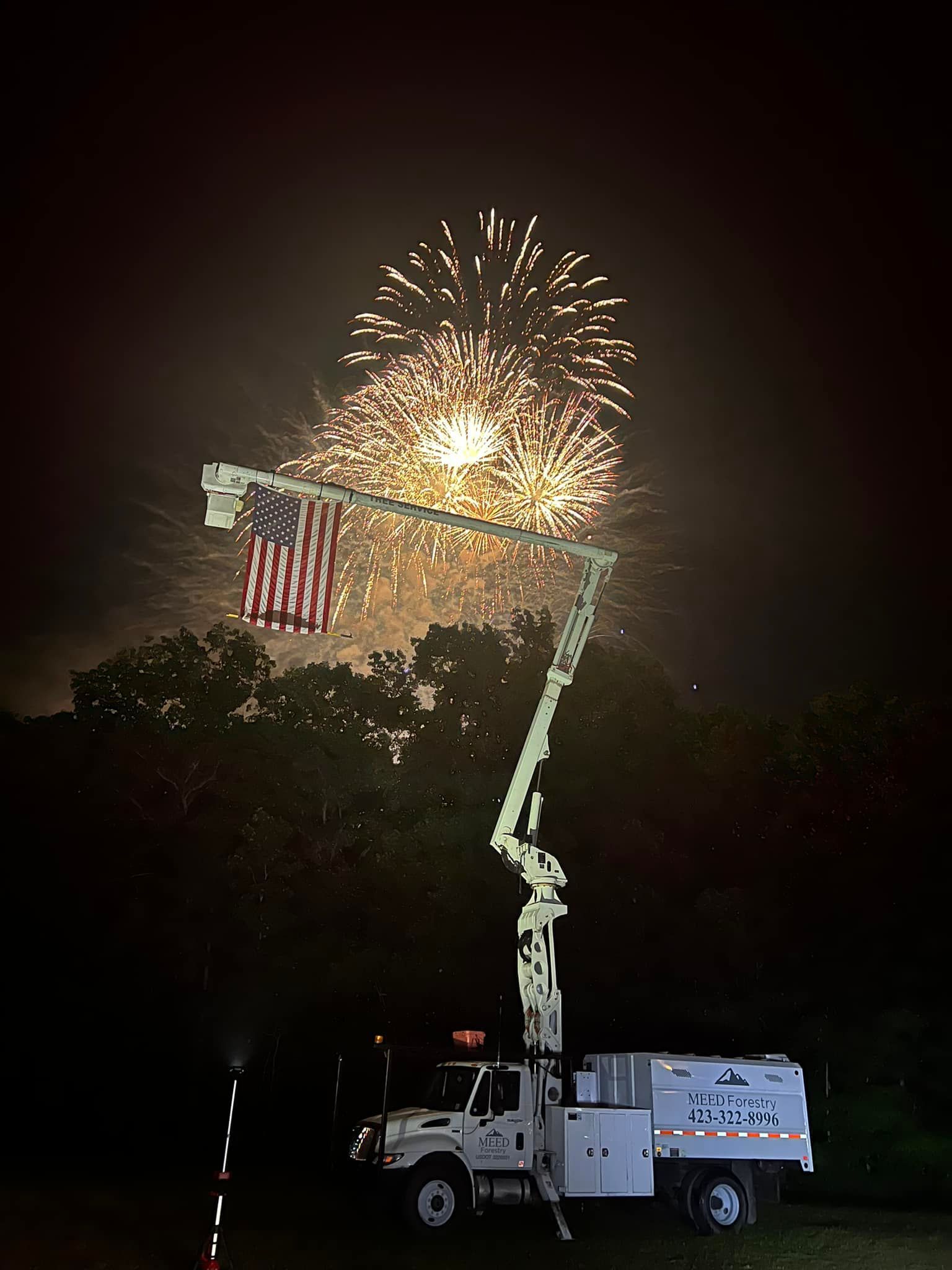 A utility bucket truck displaying an American flag with a large fireworks display in the night sky above.