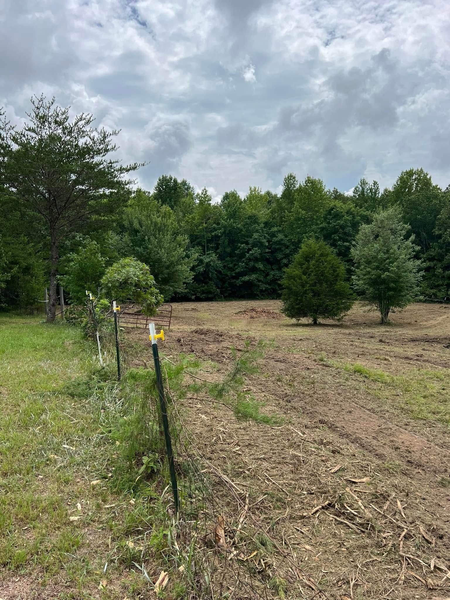 A fence line with metal posts separates a grassy area from a field of tilled soil, with a forest in the background.