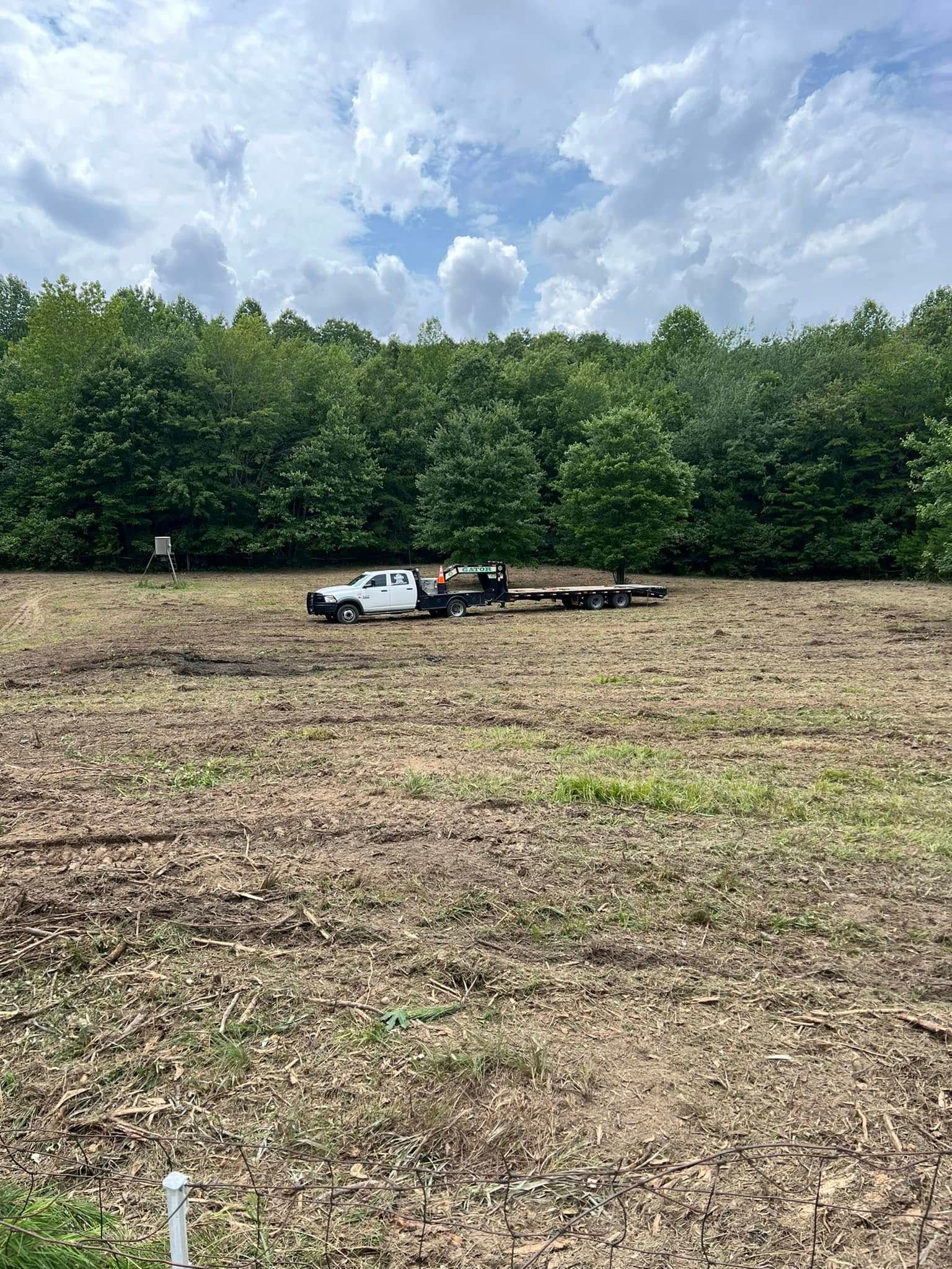 A white truck hauling a flatbed trailer sits in the middle of a large, cleared field in front of a dense tree line.