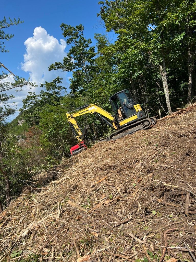 A yellow excavator with a mulcher attachment works on a steep, cleared hillside under a blue sky with scattered clouds.