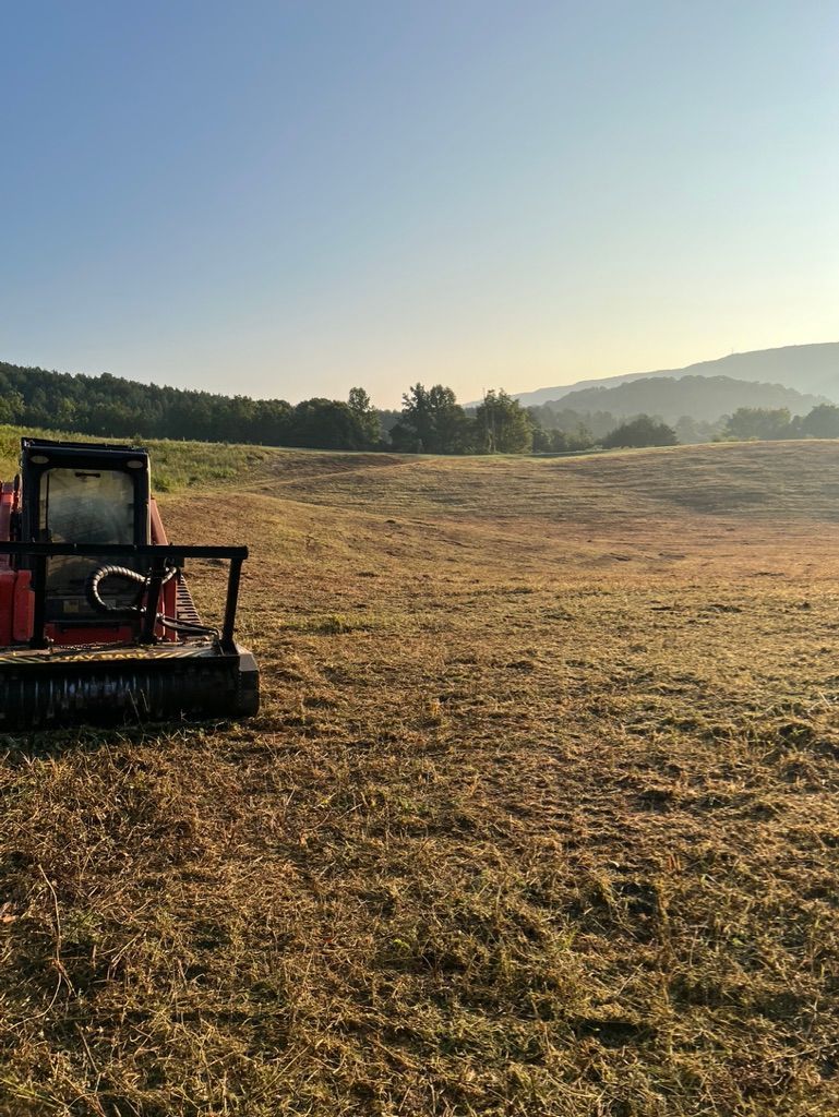 A red skid steer mower sits in a large, golden, sunlit field with rolling hills and trees in the distance.