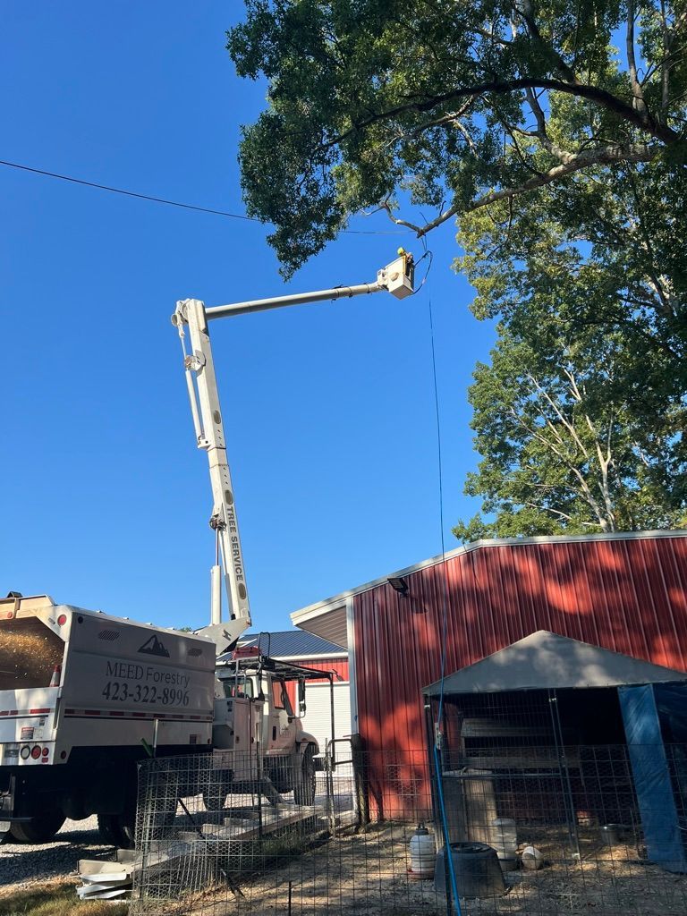 A utility truck with a raised boom and bucket is positioned next to a red barn while a worker services overhead lines.