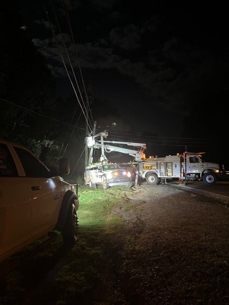 Utility trucks at night with their booms extended to work on power lines above a vehicle near a grassy roadside.