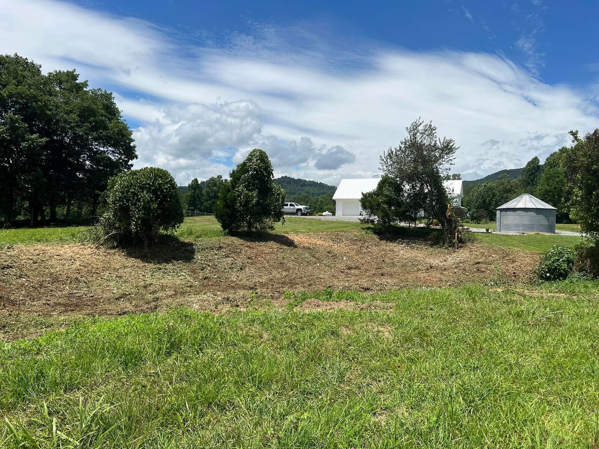 A grassy field leads up to a small rise with trees, a white building, a parked vehicle, and a metal silo under blue sky.