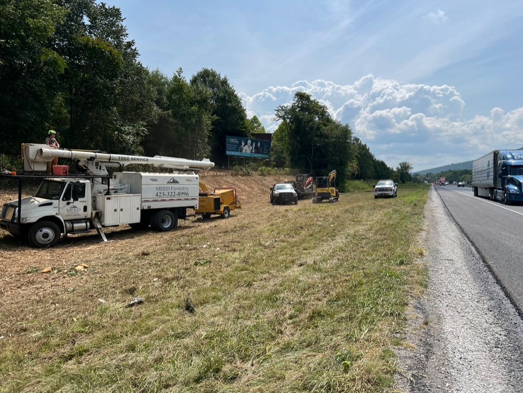 A utility bucket truck and a wood chipper parked on the grassy shoulder of a highway near a wooded area.