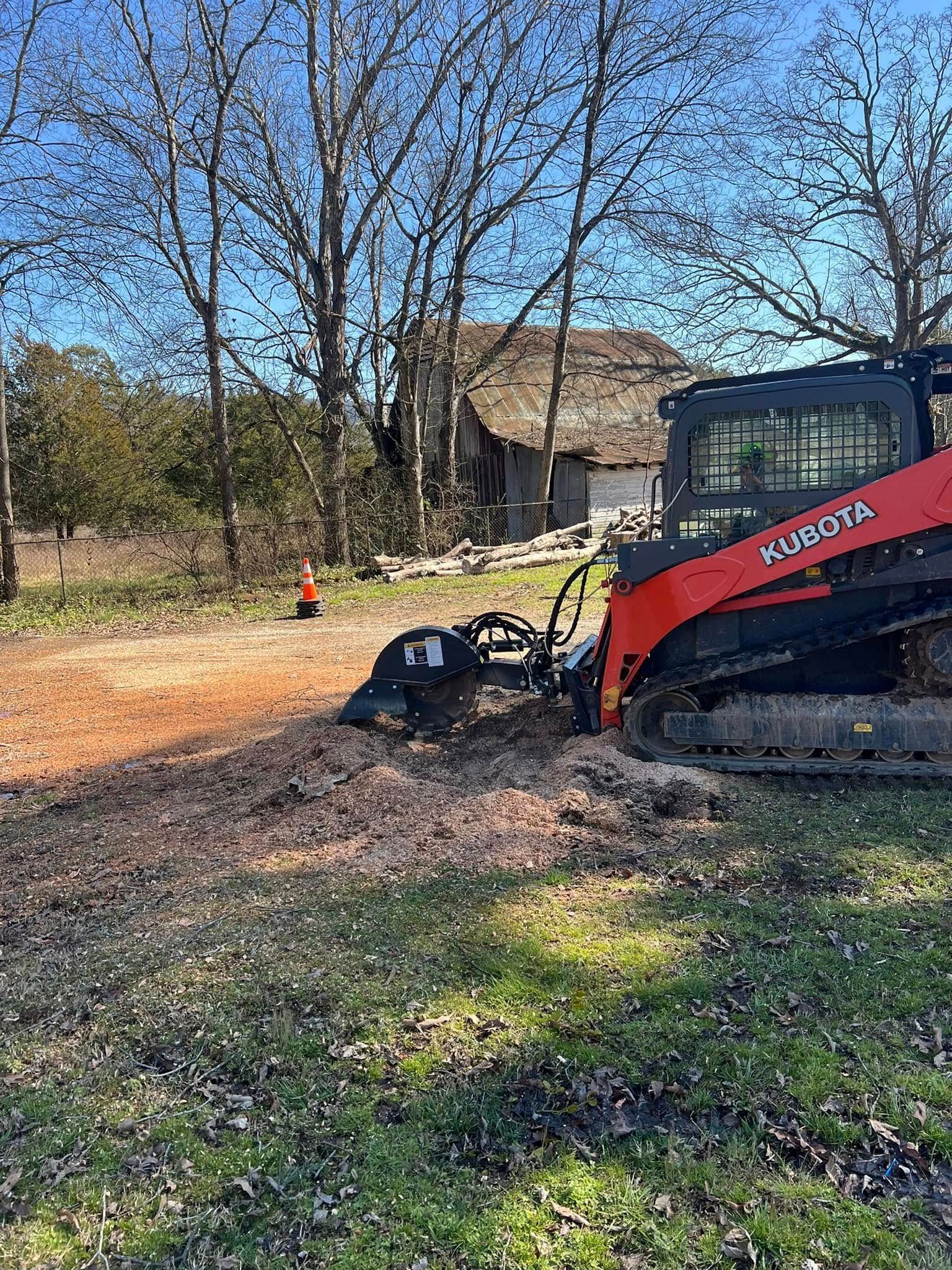 A red Kubota skid steer with a stump grinder attachment works on cleared land near a rustic building and bare trees.