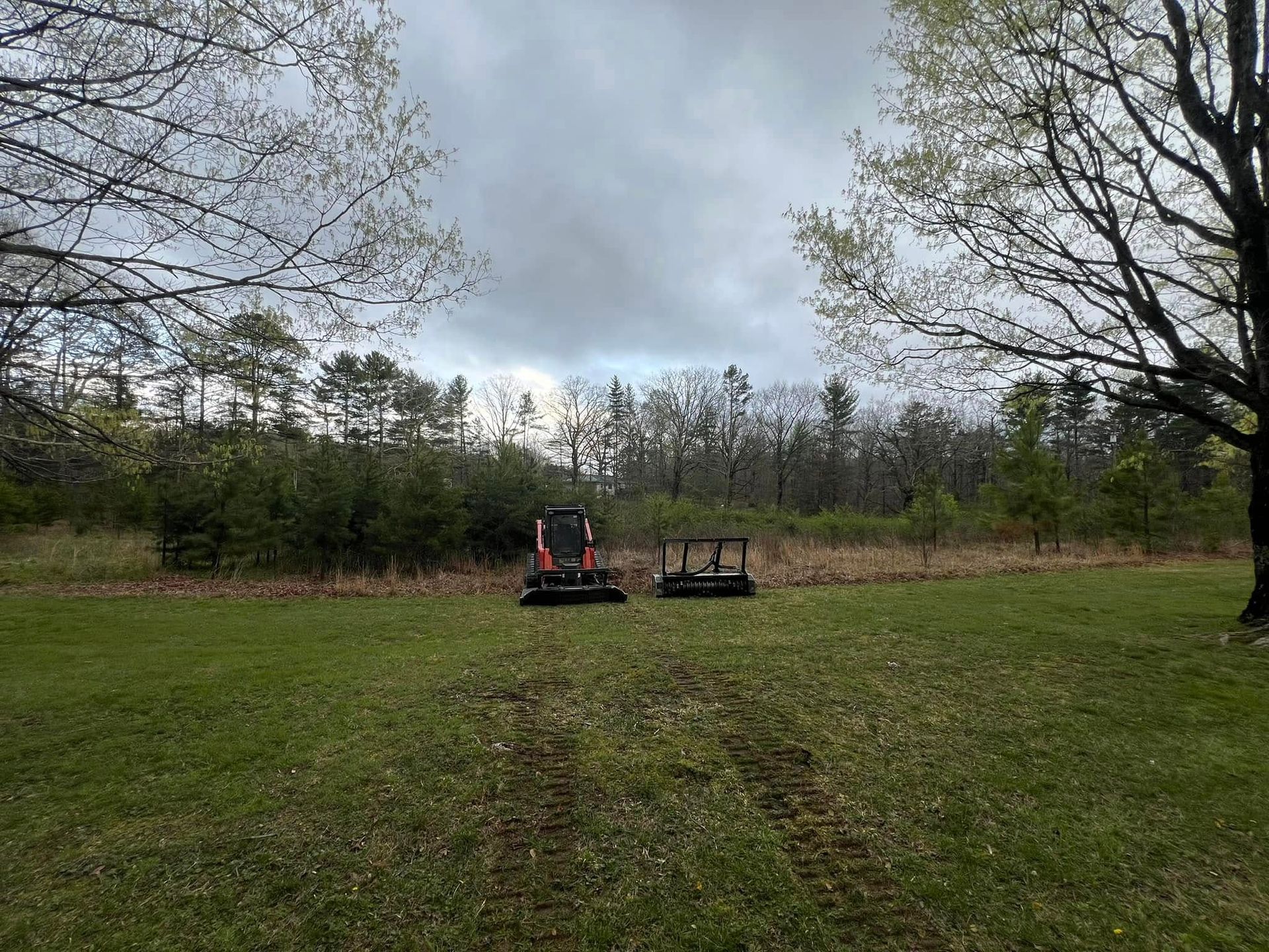 A red compact track loader and a mower parked in a grassy field before a line of trees under a cloudy sky.