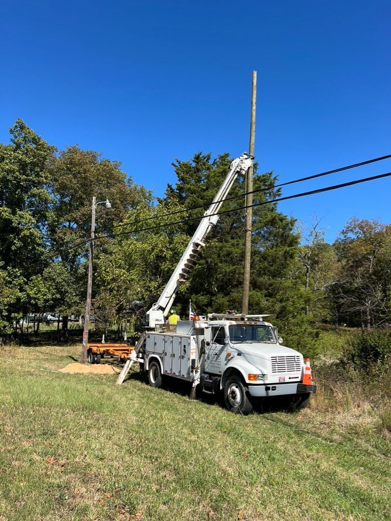 A white utility truck with an extended boom crane working on a wooden power pole in a grassy, sunlit field.
