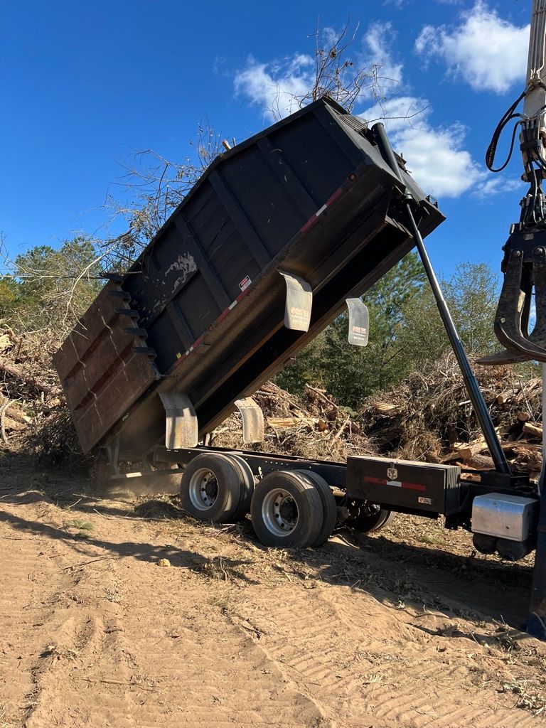 A large, black industrial dump trailer is elevated at an angle, dumping yard debris outdoors under a bright blue sky.