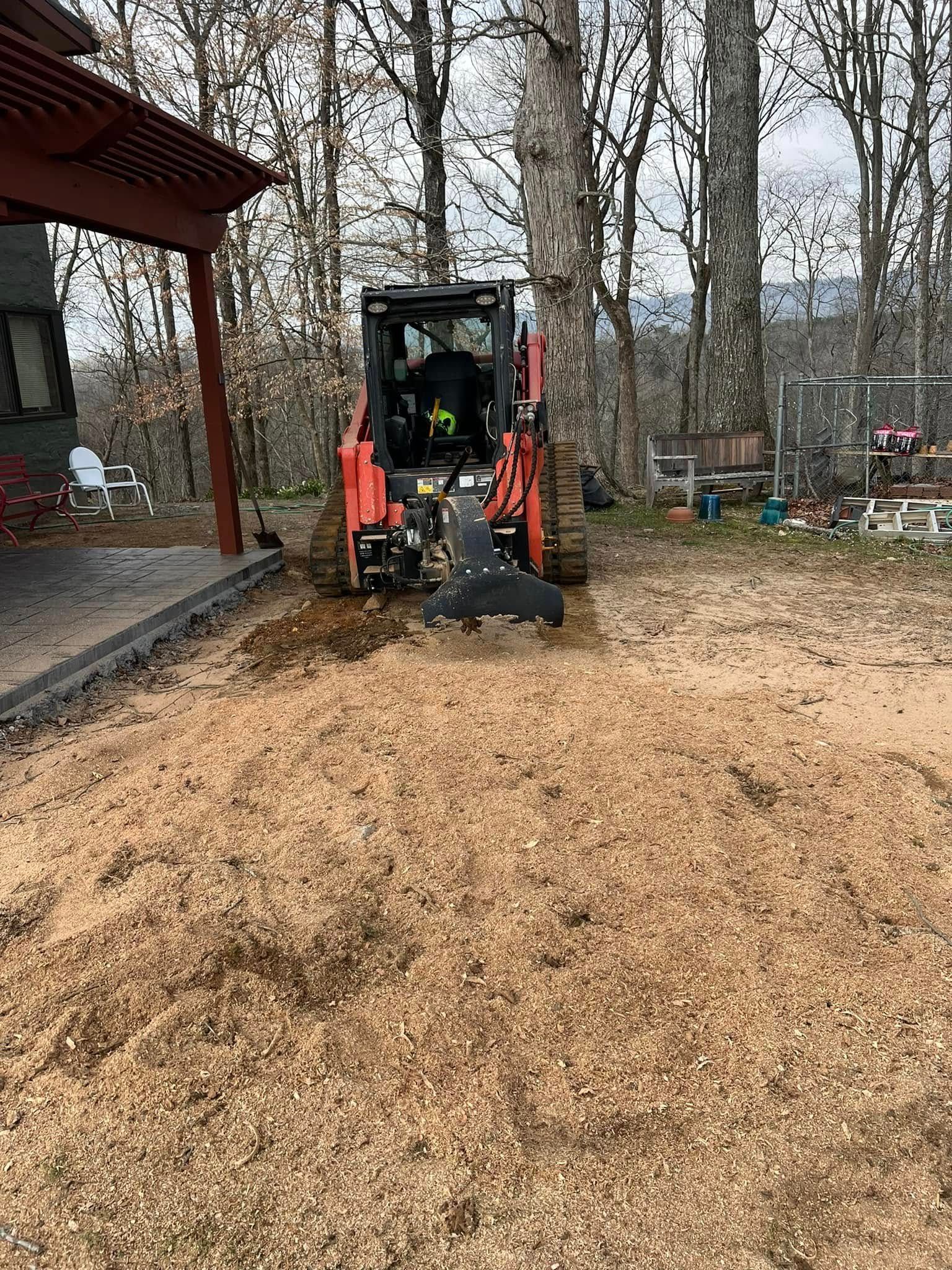 A red skid-steer loader with a stump grinder attachment works on tree debris in a yard next to a house porch.