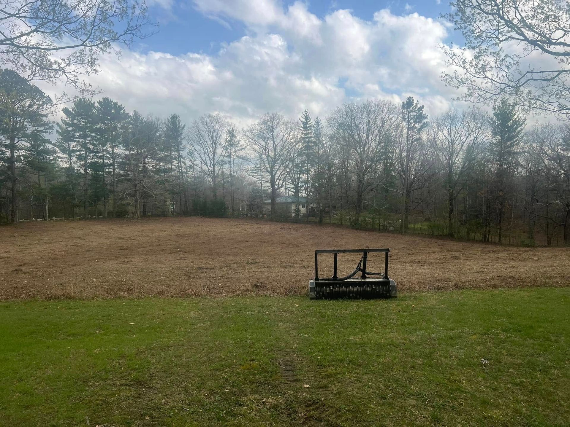 A field of dry grass and trees under a cloudy sky, with a dark metal farm implement sitting in the middle.