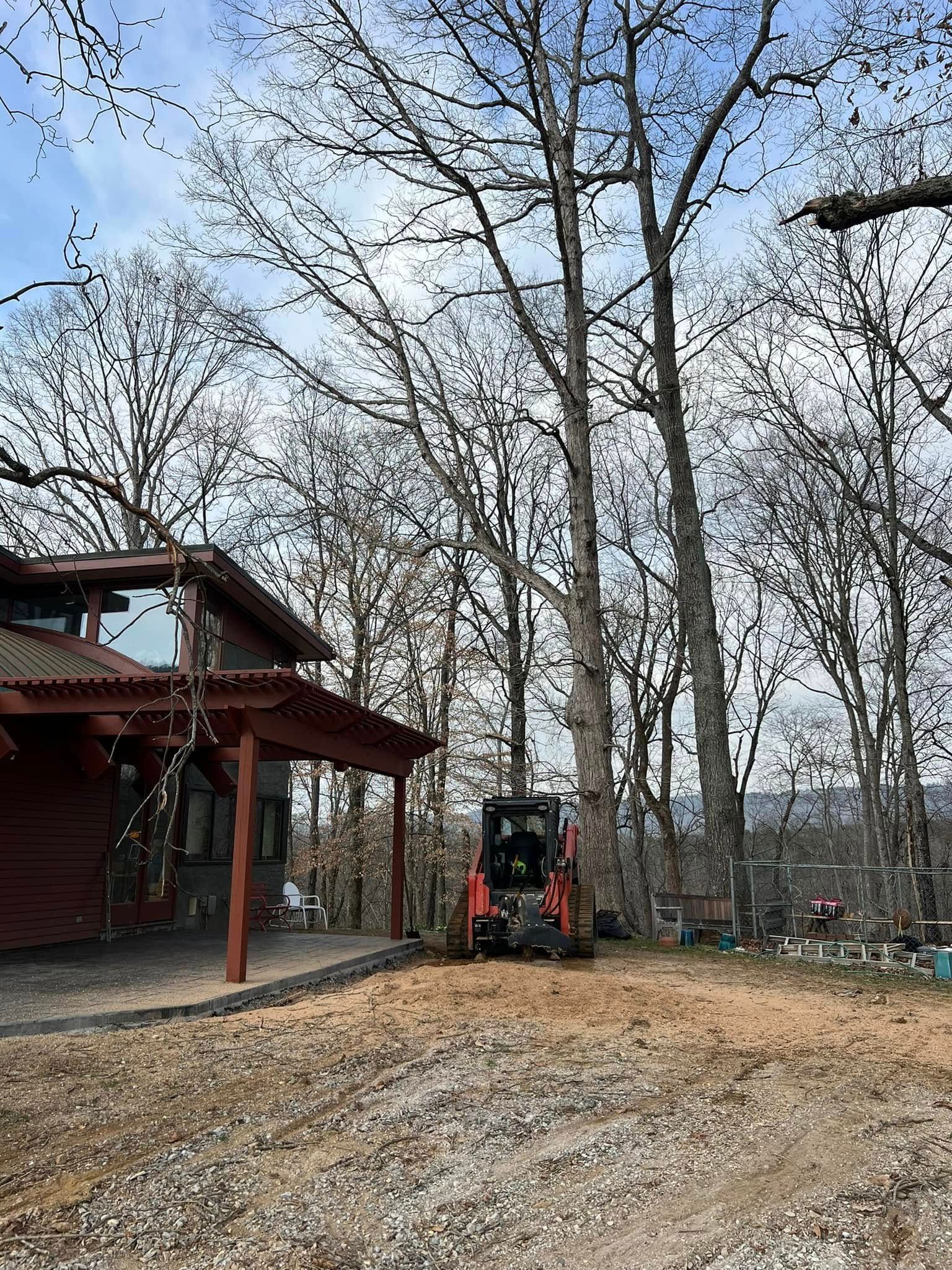 A skid steer loader sits on a gravel lot next to a wooden cabin with a porch under leafless trees on a sunny day.