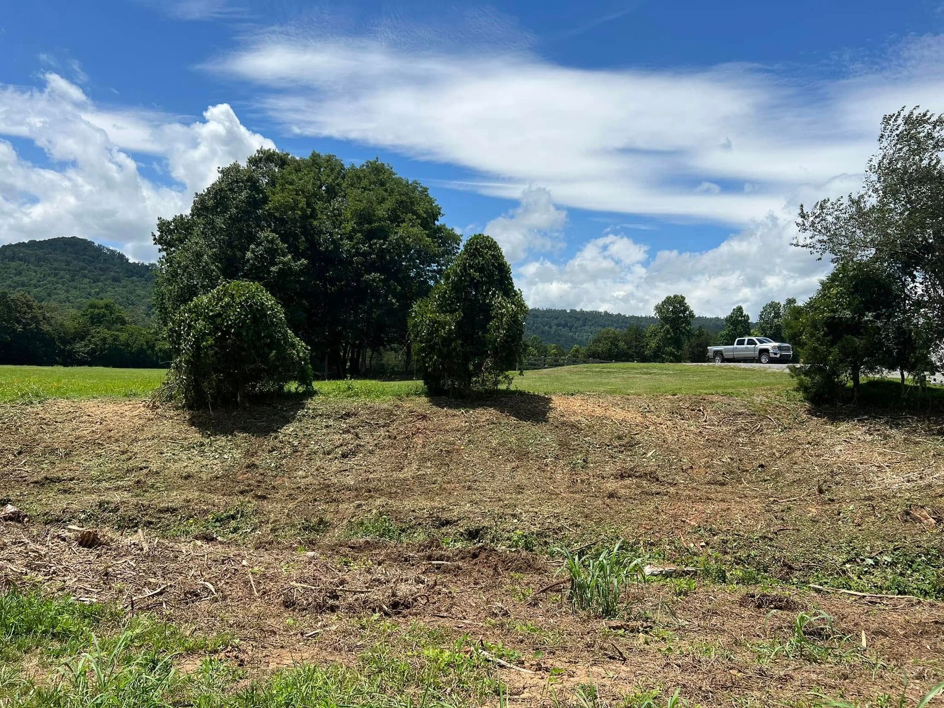 A dirt embankment leads up to a grassy field with trees, distant hills, and a white pickup truck under a blue sky.