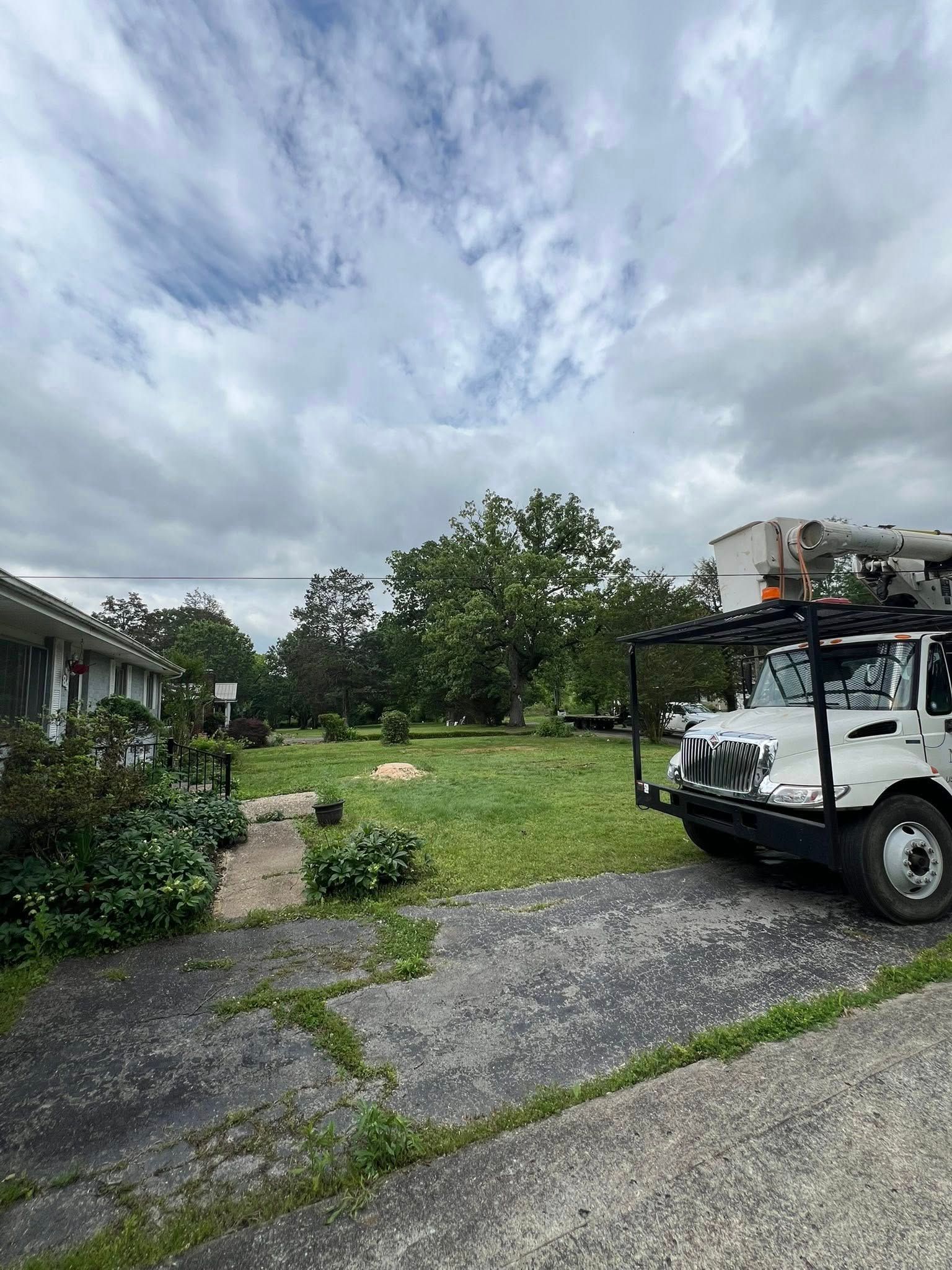 A white bucket truck is parked on a paved driveway next to a residential building and an overgrown grassy yard.