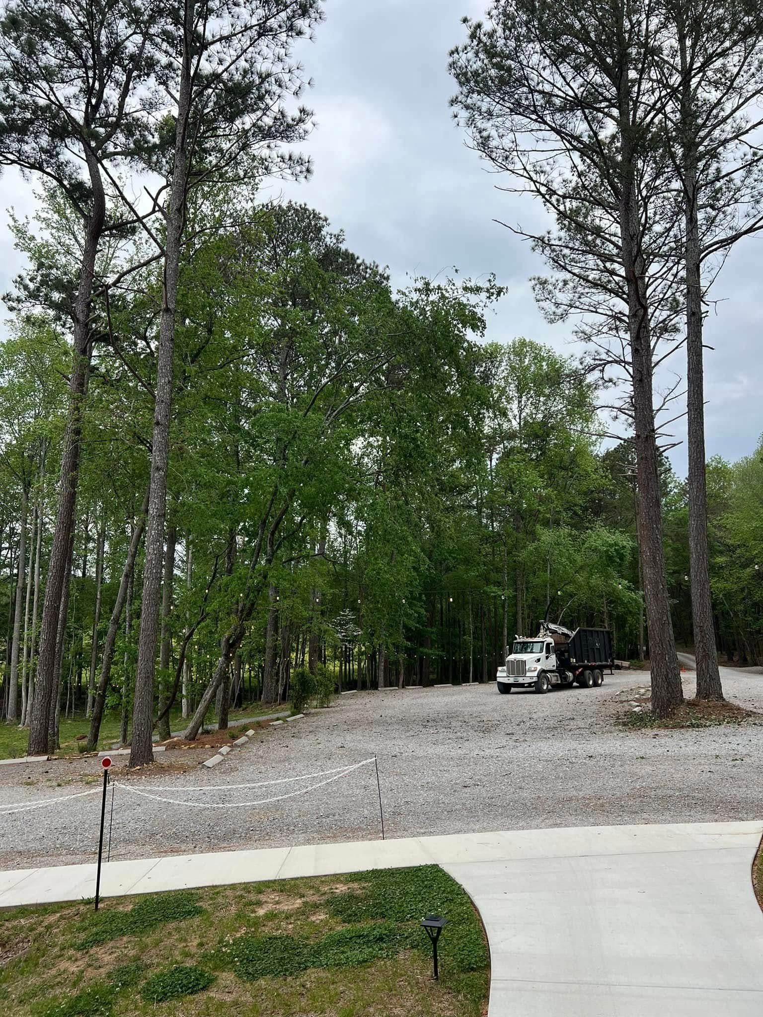 A white dump truck sits on a large gravel driveway bordered by trees and a concrete walkway in a wooded, outdoor setting.