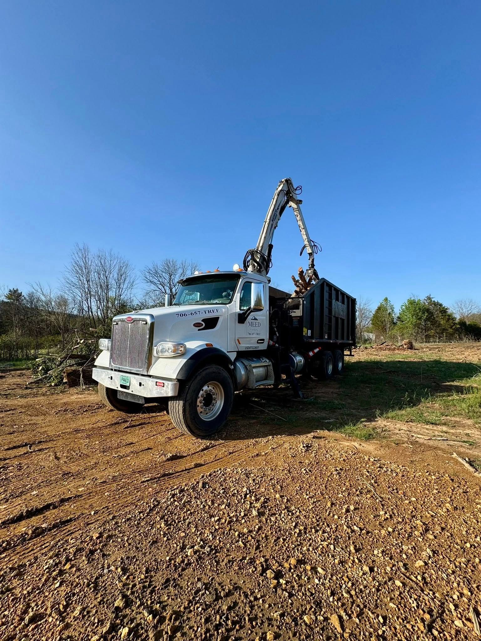 A white utility truck with a crane attachment sits in a dirt clearing against a clear blue sky.