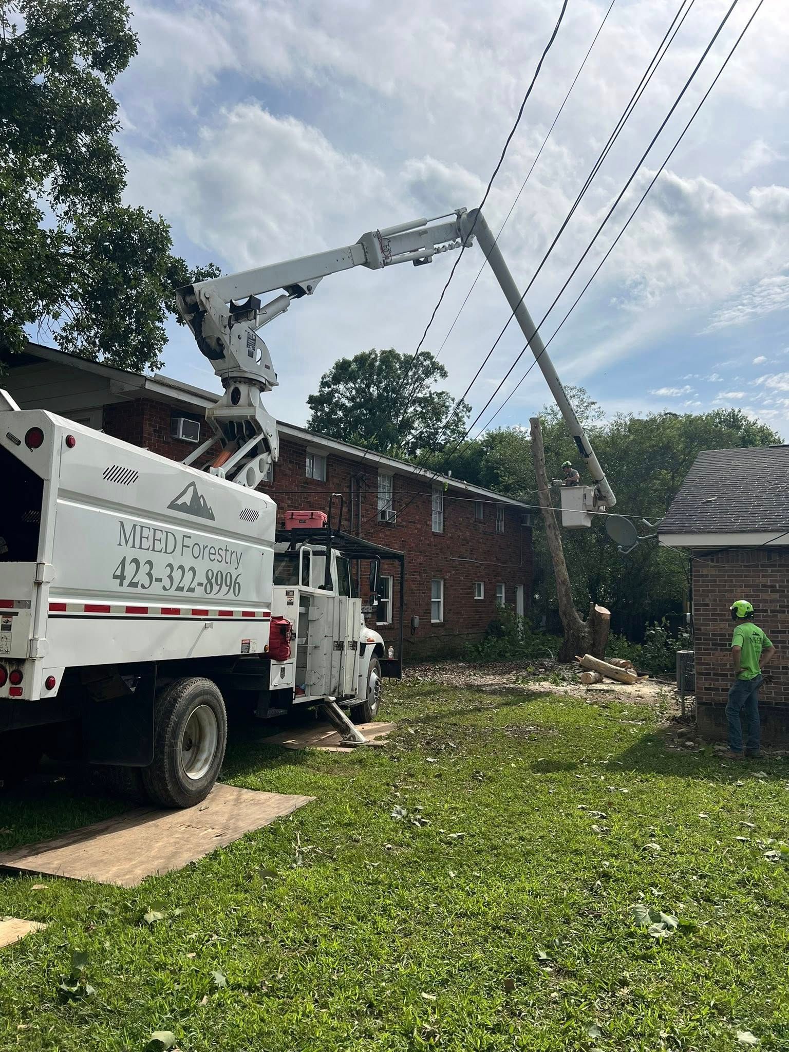 A utility truck with an extended boom bucket parked near a brick building, with a worker standing on the ground nearby.