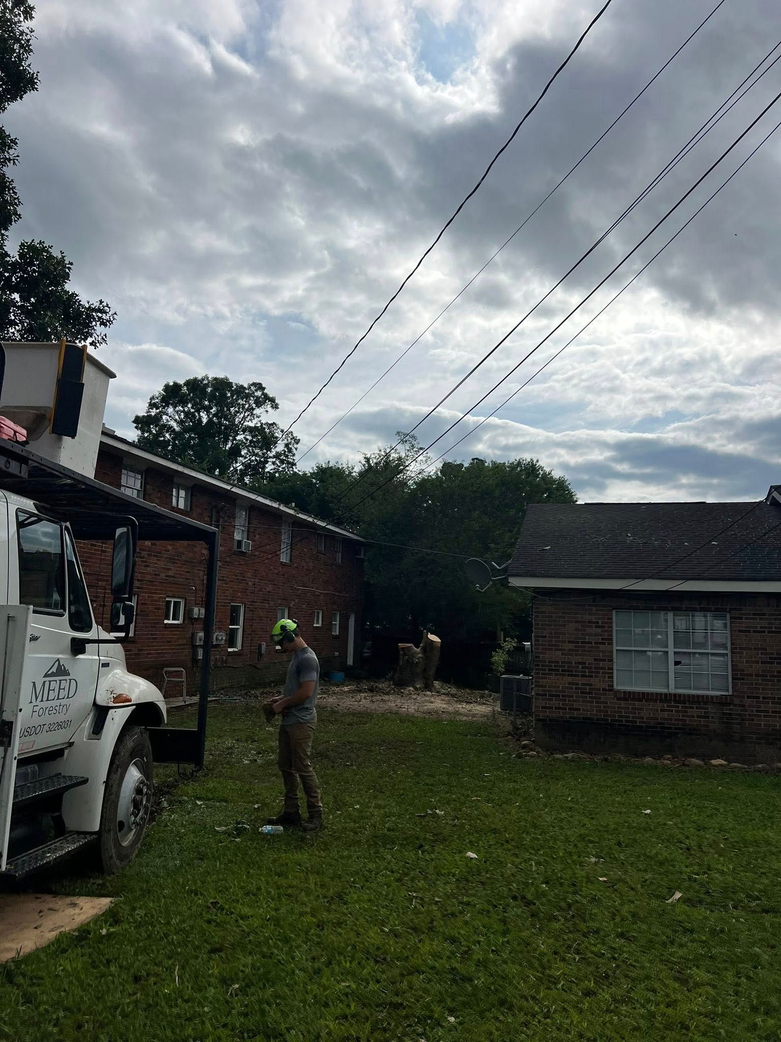 A worker in a helmet stands near a utility truck parked in a grassy yard beside brick buildings under a cloudy sky.