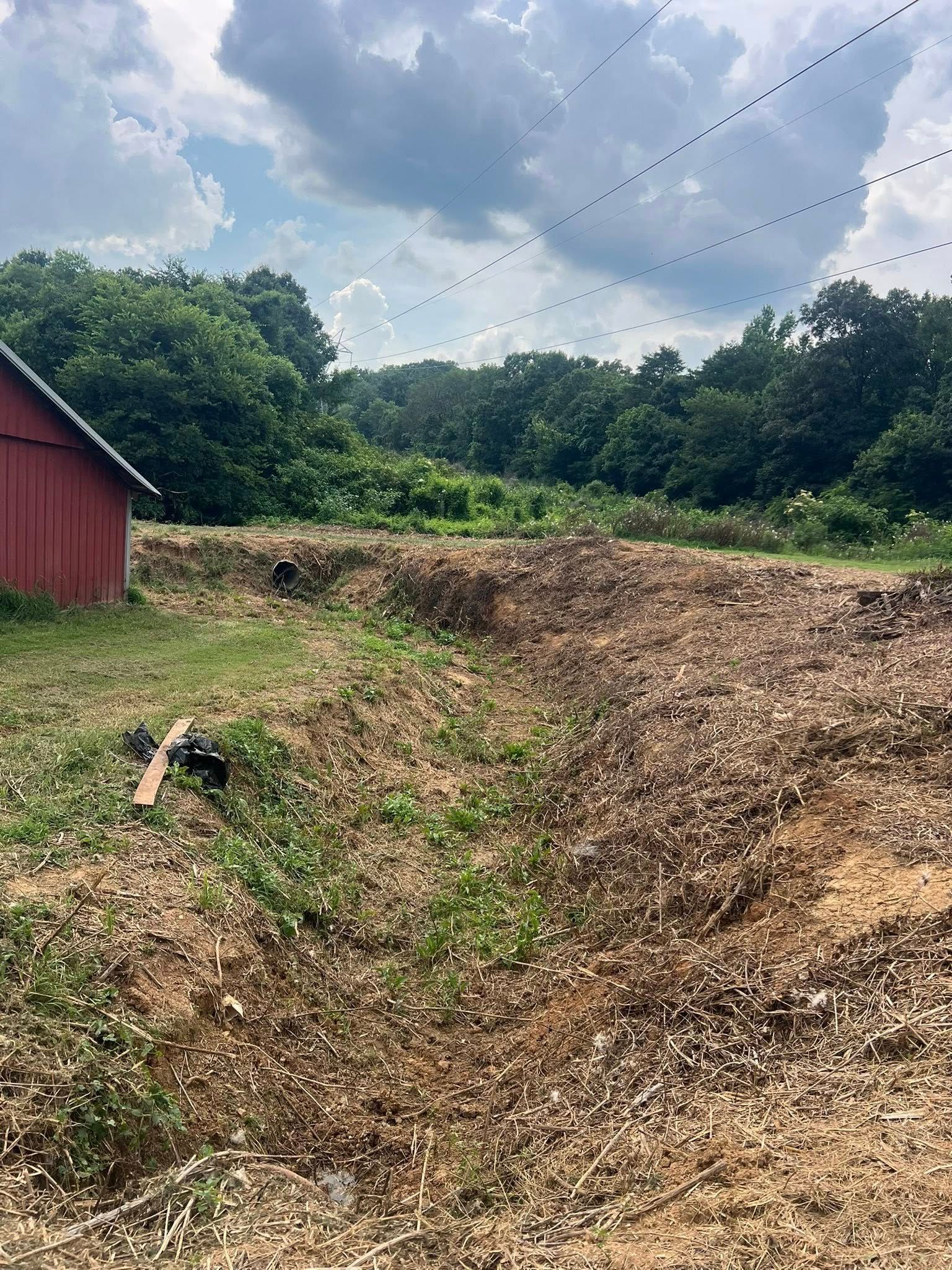 A red barn stands beside a patch of cleared, uneven earth and brush in a rural area under a cloudy sky.